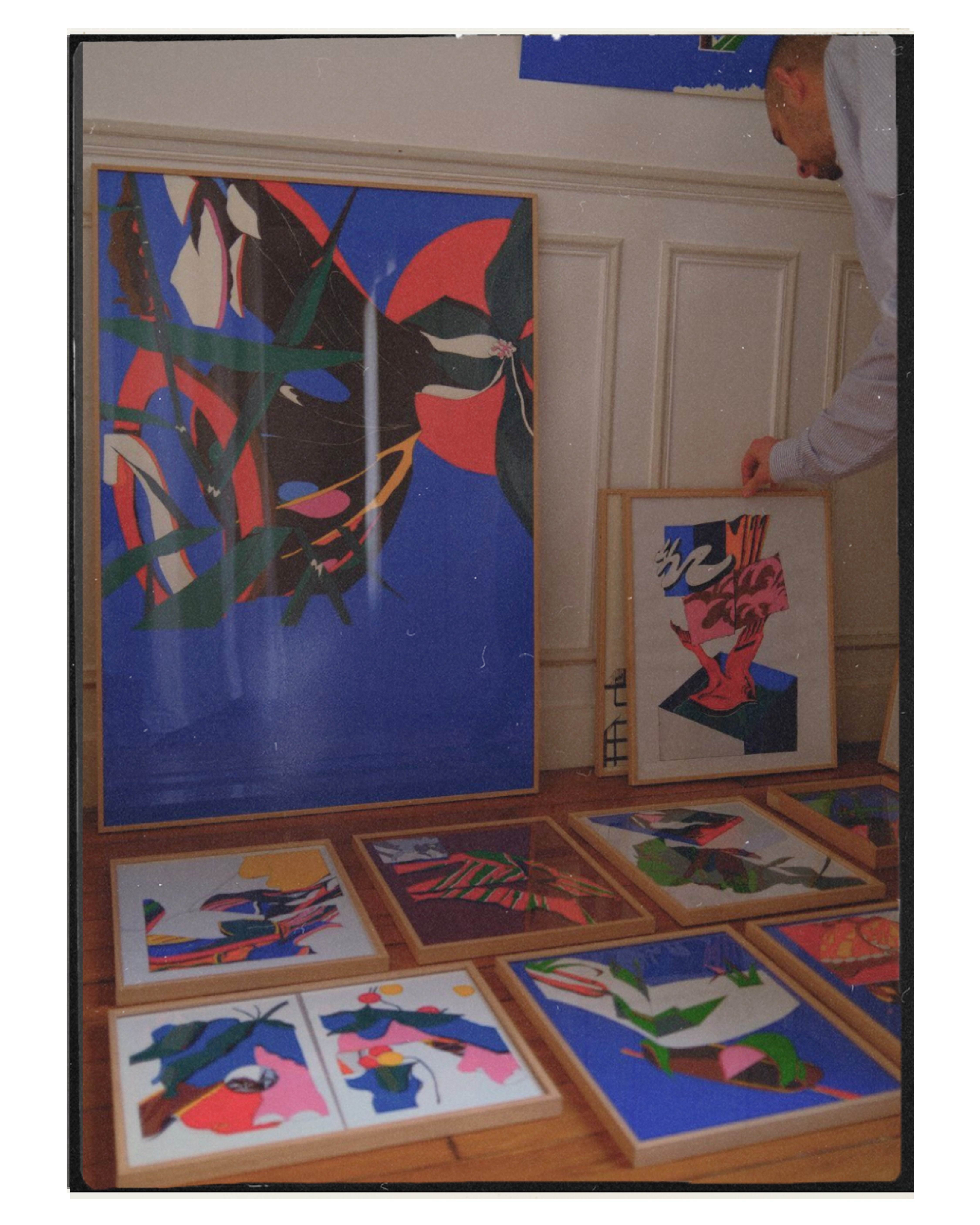 A person arranging several abstract and colorful framed artworks on a wooden floor, featuring bold shapes and dynamic compositions against a neutral wall background.