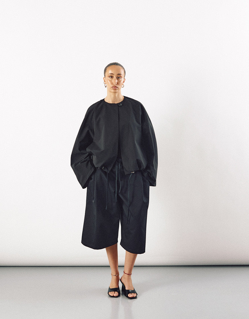 Model wearing a black minimalist jacket with a round neckline and voluminous sleeves paired with loose, knee-length black shorts, highlighting understated elegance and subtle pocket details.
