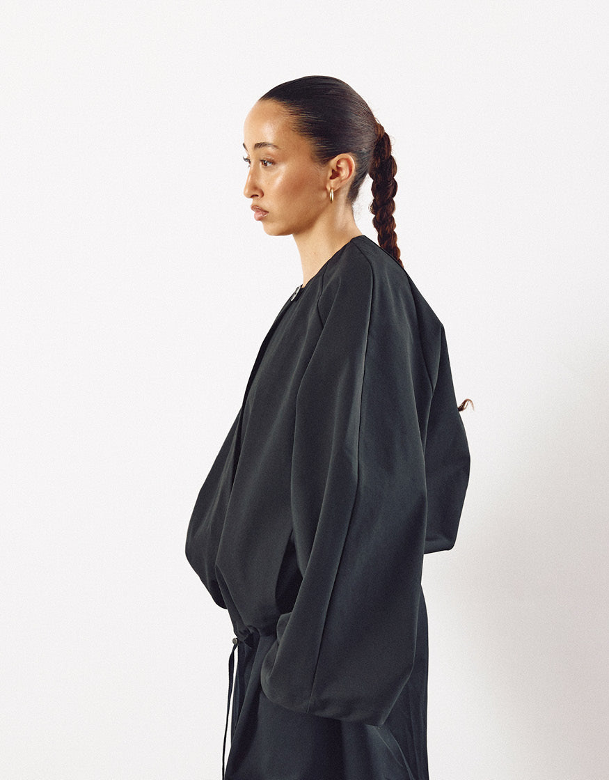 Side profile of a model wearing a minimalist black jacket with oversized sleeves and discreet pockets, showcasing understated elegance and contemporary design.