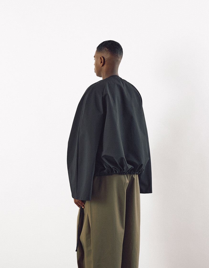Rear view of a model wearing a minimalist black jacket with subtle volume and elastic detail at the hem, paired with loose olive trousers, emphasizing understated elegance.