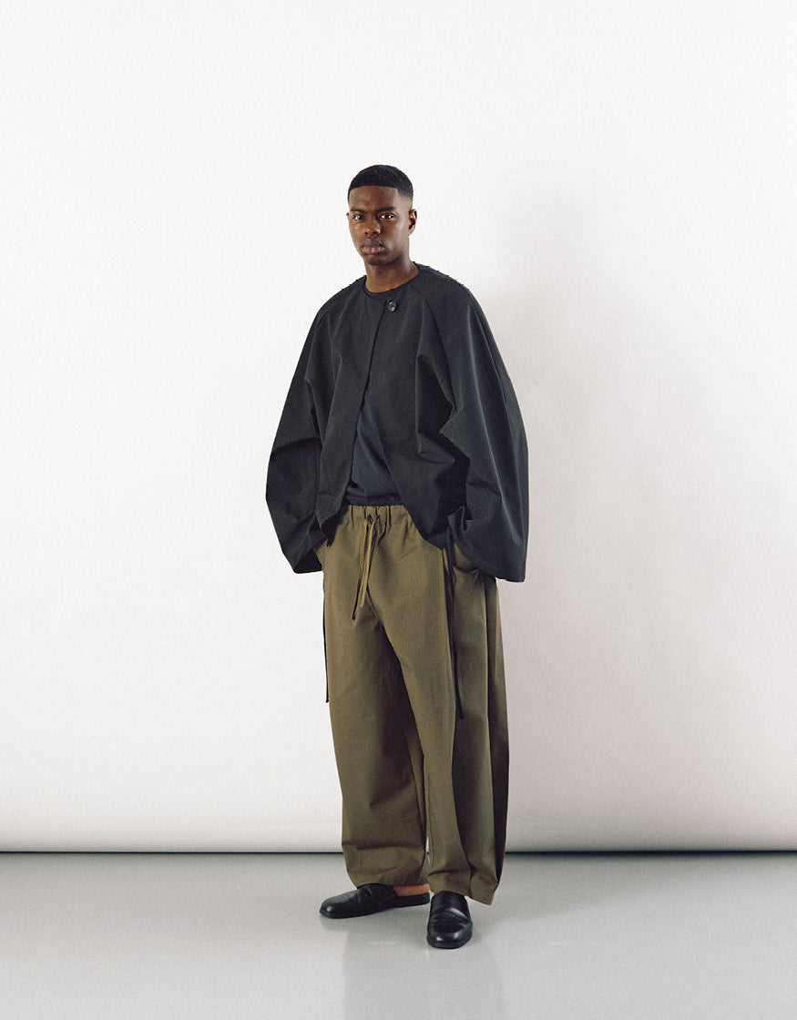 Man wearing a black minimalist jacket with pockets paired with loose olive trousers and black shoes, showcasing understated, contemporary unisex outerwear design.