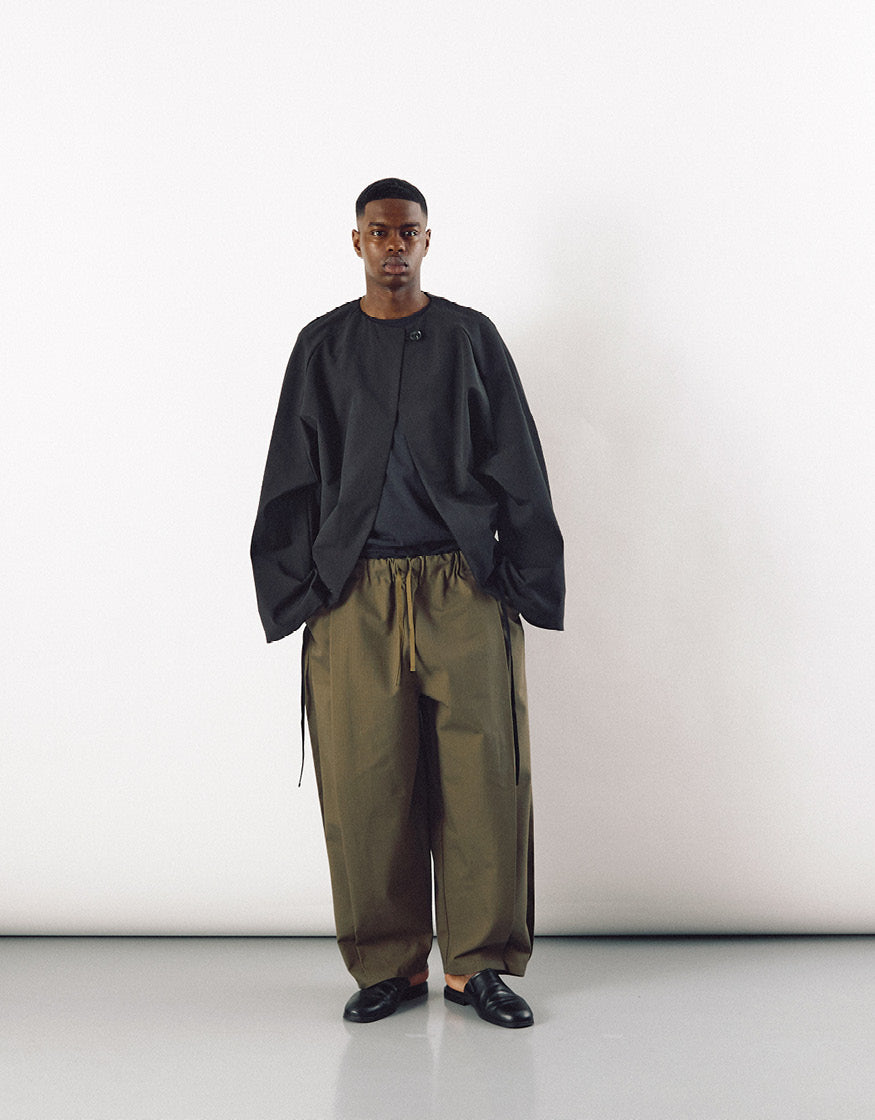 Male model wearing a minimalist black jacket with pockets, featuring a relaxed fit and understated design, paired with loose olive-green trousers and black slip-on shoes.