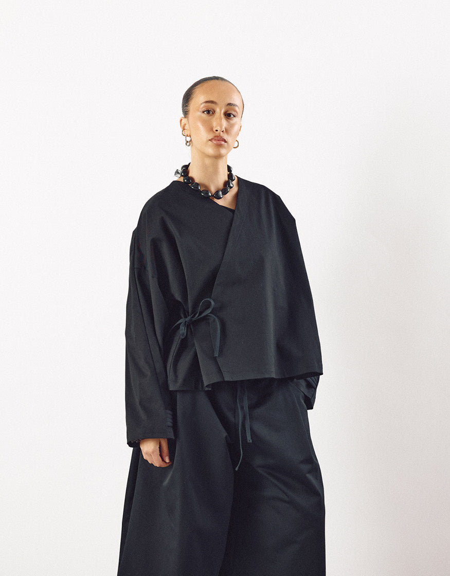 Model wearing a minimalist black kimono-style top with asymmetrical design and loose sleeves, paired with matching wide-leg pants, showcasing understated elegance and volume.