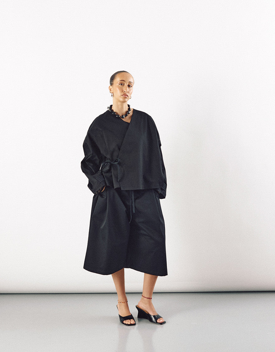 Model wearing an elegant black minimalist kimono-inspired top with wide sleeves and matching wide-leg cropped pants, styled with a statement necklace and open-toe heels.