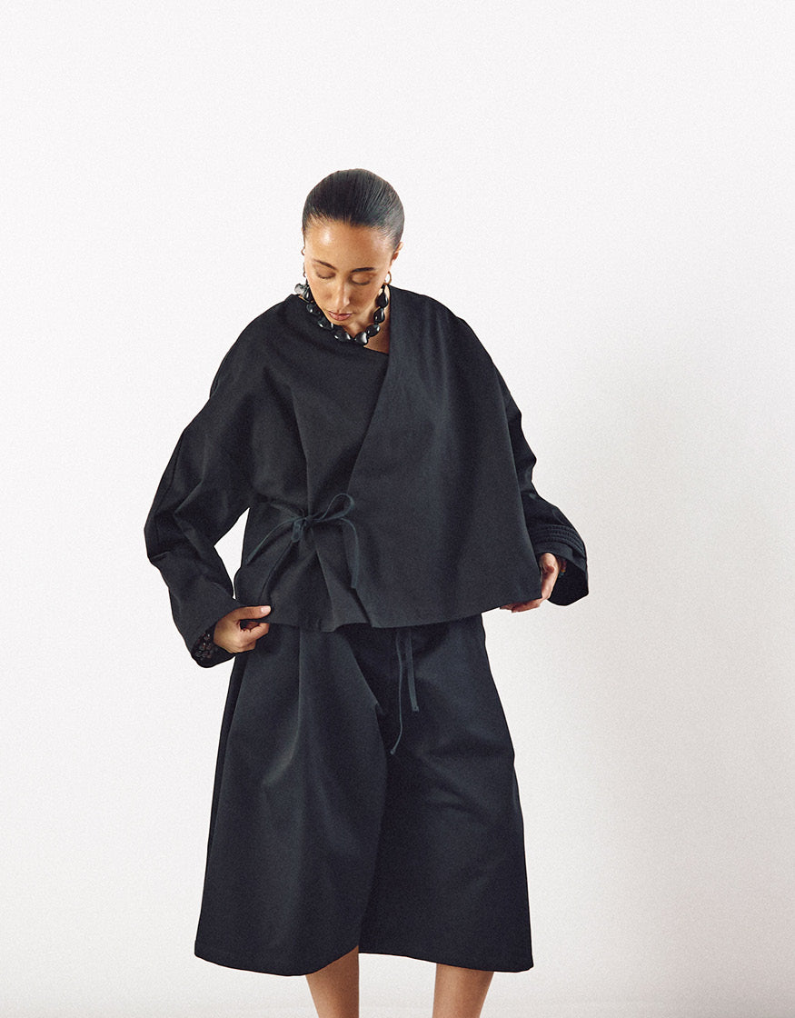 Woman wearing a minimalist black asymmetrical kimono-style top with wide sleeves and loose black culottes, showcasing understated elegance and contemporary design.