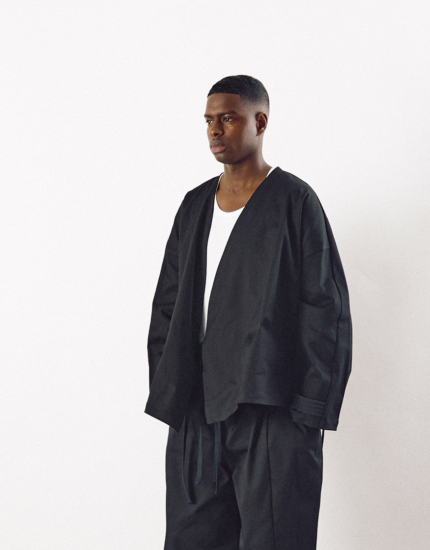 Male model wearing a minimalist black oversized jacket with dropped shoulders and loose tailored pants, paired with a simple white shirt for a contemporary, elegant look.