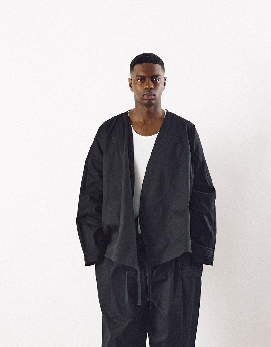 A model wearing a minimalist black asymmetrical jacket over a white top paired with loose black pants, showcasing a contemporary unisex design with clean lines and volume.