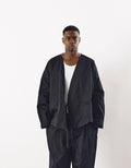 A model wearing a minimalist black asymmetrical jacket over a white top paired with loose black pants, showcasing a contemporary unisex design with clean lines and volume.