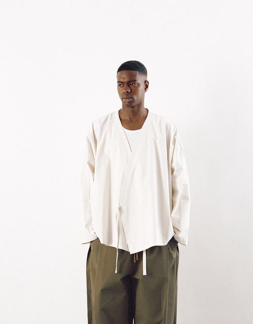Male model wearing an off-white minimalist kimono-style top with relaxed fit and subtle asymmetrical design, paired with loose olive green trousers.