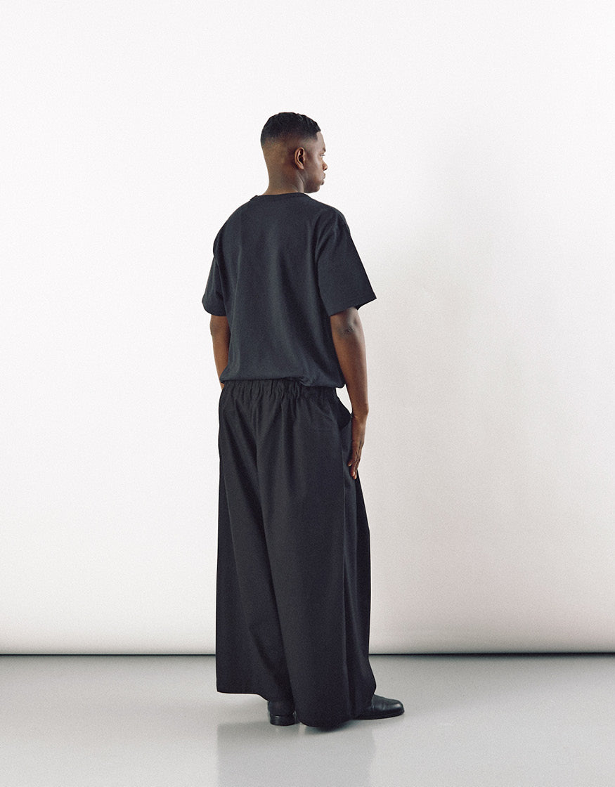 A man wearing black wide-leg pants paired with a loose black top, standing against a plain light background, showcasing minimalist unisex contemporary fashion.