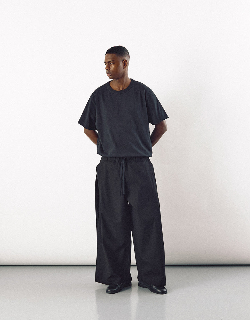 Man wearing black wide pants paired with a loose black short-sleeve shirt, standing against a plain white background in a minimalist, contemporary style.