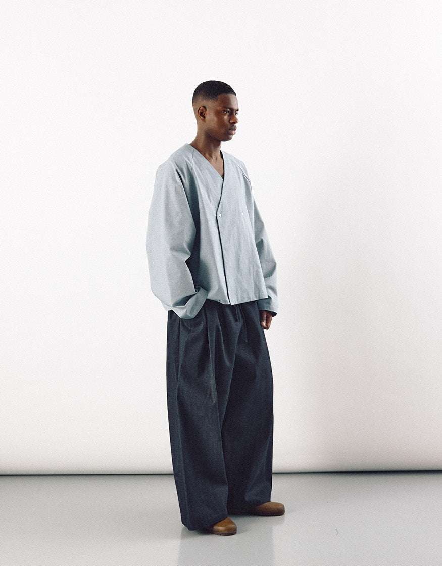 A male model wearing loose-fitting indigo wide pants paired with a light gray oversized jacket, showcasing minimalist, unisex fashion with a focus on volume and understated elegance.