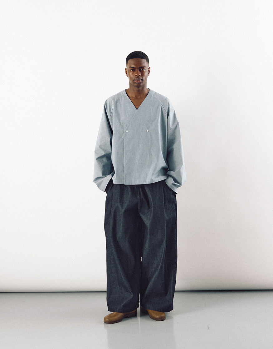 Man standing against plain background wearing a minimalist long-sleeve V-neck top paired with wide indigo pants featuring a relaxed silhouette emphasizing volume and comfort.