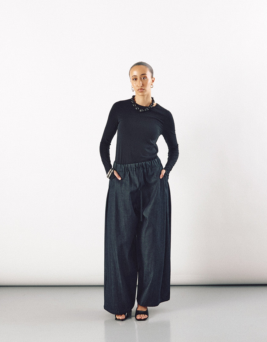 Woman wearing a minimalist outfit featuring wide indigo pants with an elastic waistband, paired with a fitted black long-sleeve top and black open-toe sandals, standing against a plain white background.