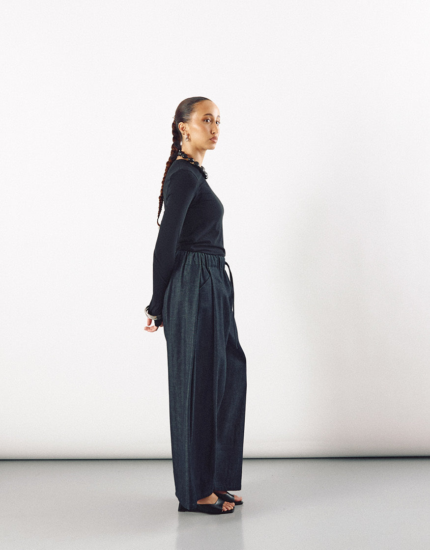 Profile view of a woman wearing minimalist wide indigo pants paired with a fitted long-sleeve black top, showcasing elegant relaxed volume and contemporary, timeless style.