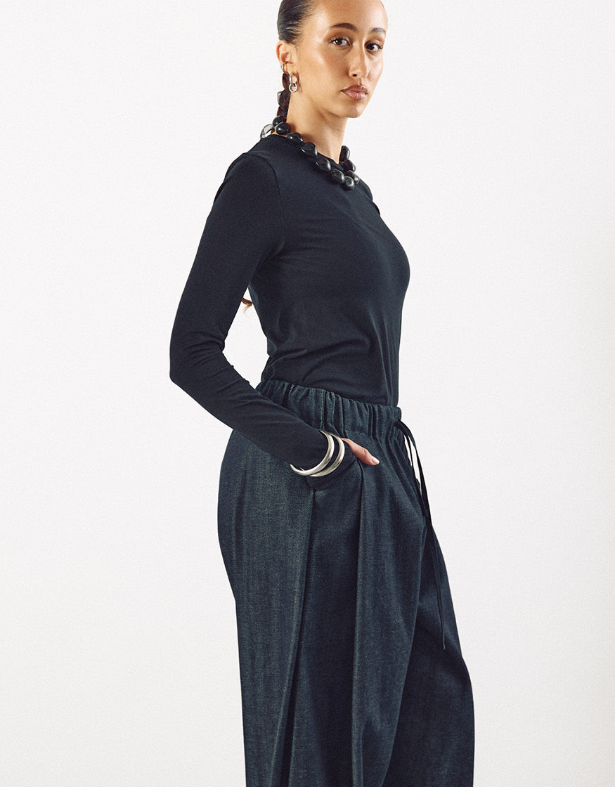 Side view of a woman wearing wide indigo pants with an elastic waistband paired with a fitted black long-sleeve top, accessorized with bold jewelry, showcasing minimalist and elegant style.
