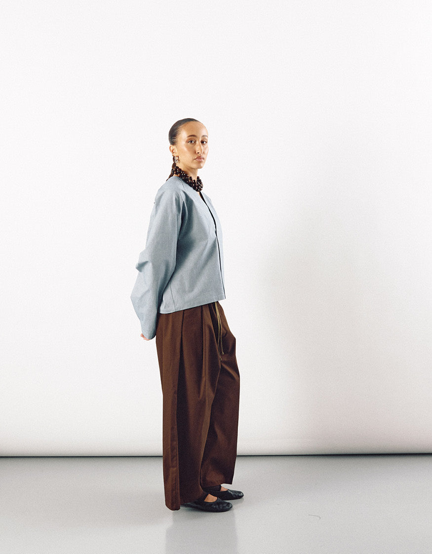 Model wearing tobacco-colored wide pants paired with a loose-fitting light gray top, showcasing minimalist, unisex design with a focus on volume and clean lines.