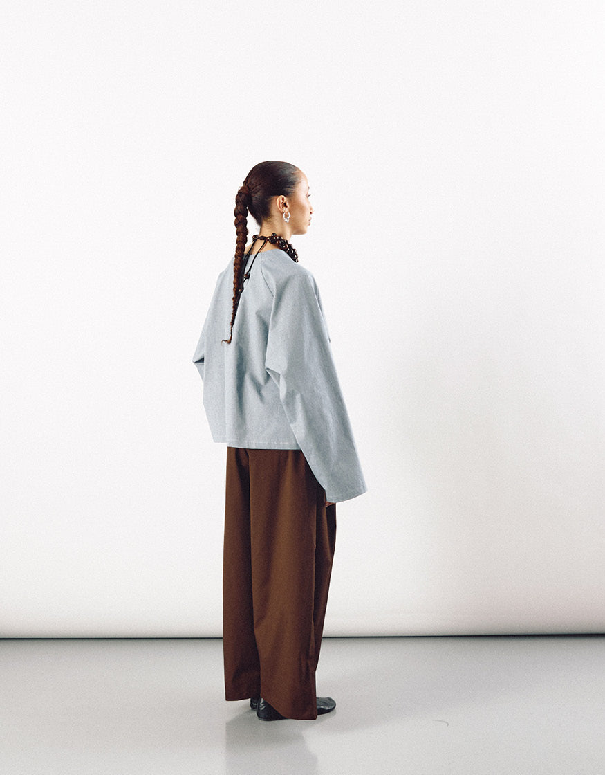 Side profile of a model wearing minimalist wide tobacco-colored pants paired with a loose, light gray top, highlighting a contemporary unisex silhouette.
