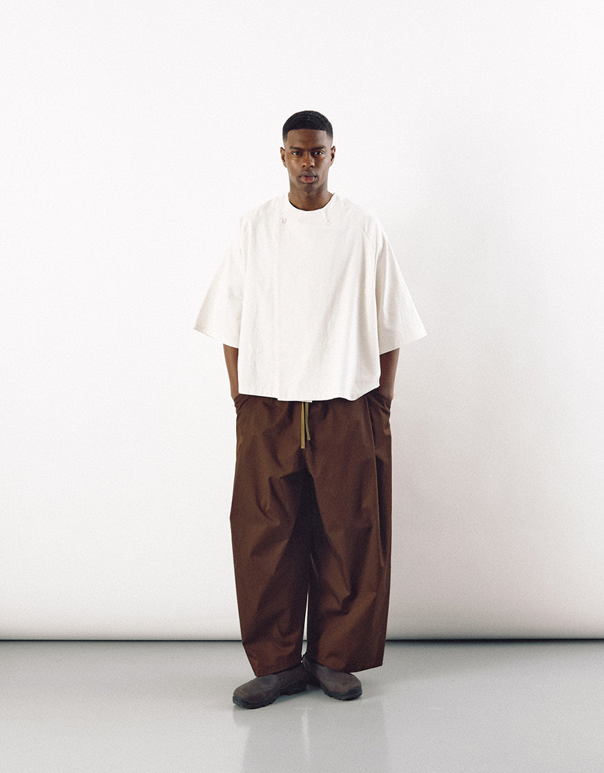 Male model wearing minimalist wide tobacco-colored pants paired with an oversized white top, showcasing a unisex, contemporary design focused on volume and understated elegance.