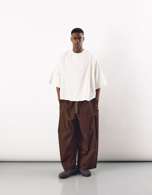 Male model wearing minimalist wide tobacco-colored pants paired with an oversized white top, showcasing a unisex, contemporary design focused on volume and understated elegance.