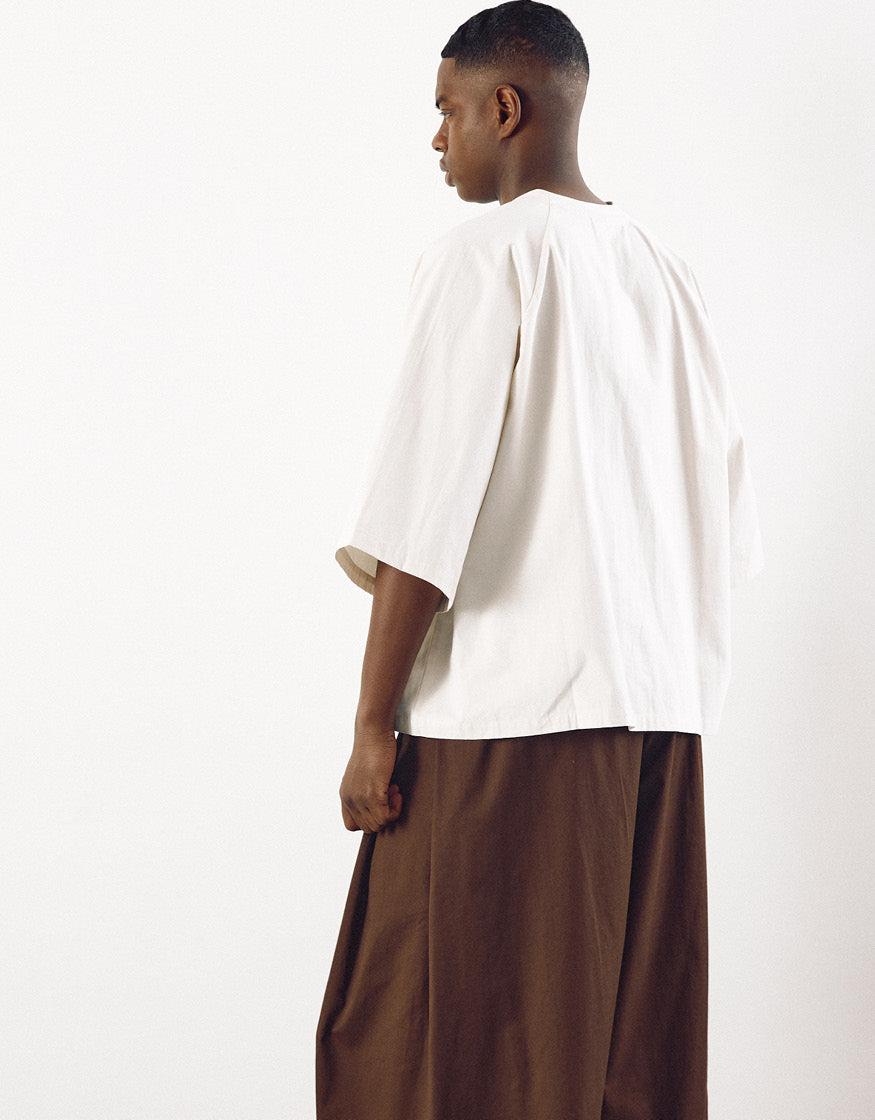 Male model seen from the back wearing wide tobacco-colored pants and an oversized white top, showcasing minimalist design and relaxed volume for freedom of movement.