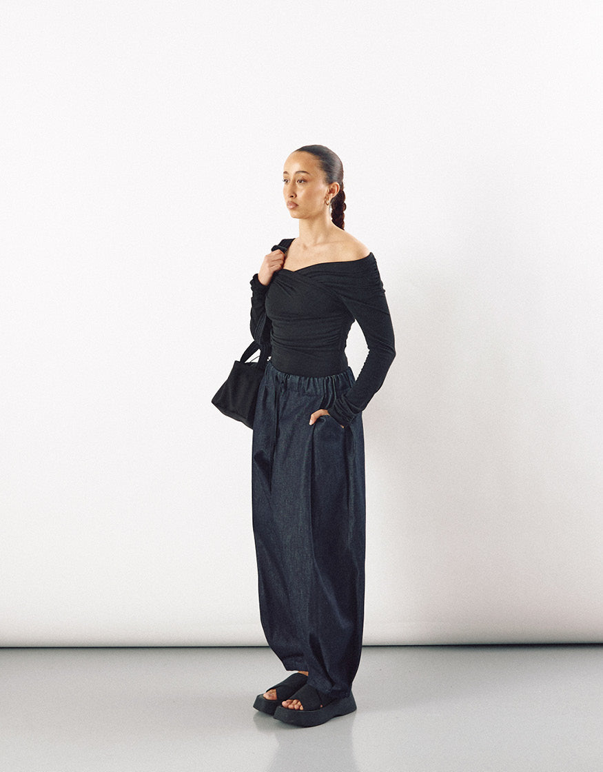 Woman wearing loose indigo pants with a gathered waist paired with an off-shoulder black top, carrying a black bag and wearing black platform sandals, showcasing minimalist and elegant style.
