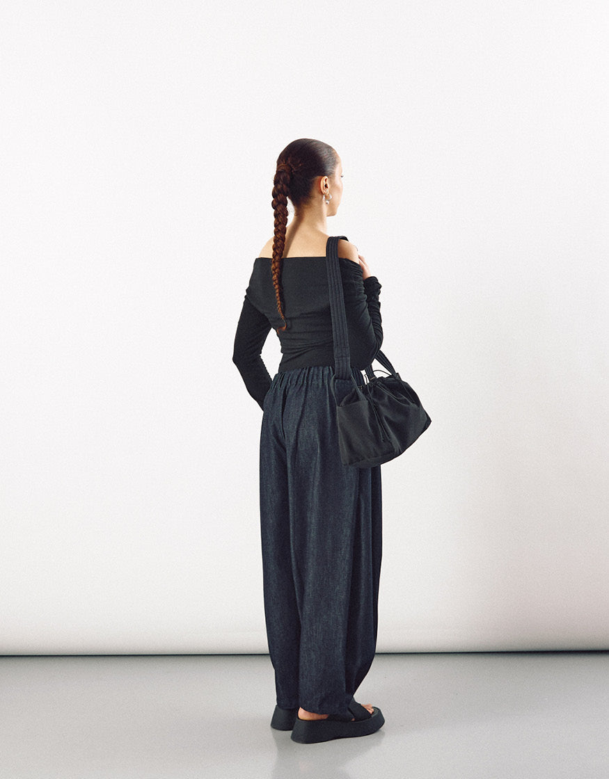 Woman seen from behind wearing loose indigo pants with an elastic waistband, a fitted off-the-shoulder black top, carrying a black shoulder bag, showcasing minimalist contemporary style.