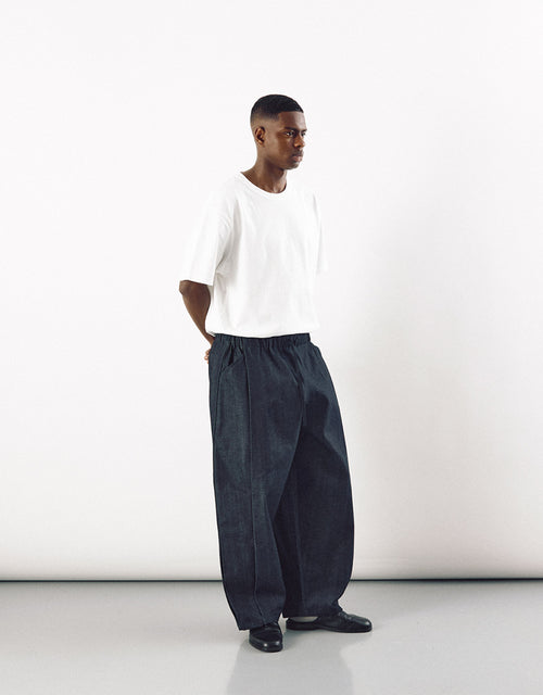 Man wearing minimalist dark indigo curved pants with a relaxed fit and a plain white short sleeve shirt, standing against a white background.