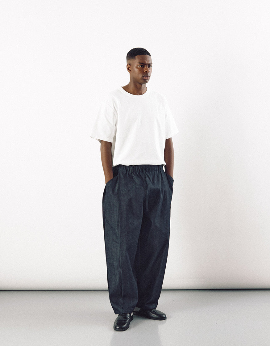 A model wearing loose-fitting indigo pants with a high waist and subtle pleats, paired with a simple white t-shirt and black shoes, highlighting minimalist unisex style.