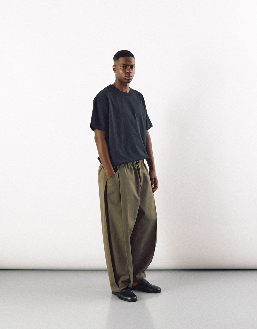 A male model stands wearing olive minimalist curved pants paired with a loose black t-shirt and black shoes, showcasing subtle volume and contemporary design.