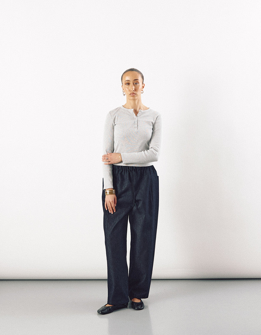 Woman wearing relaxed indigo tapered pants paired with a fitted light gray long-sleeve top, standing in a minimalist studio setting with a neutral background.