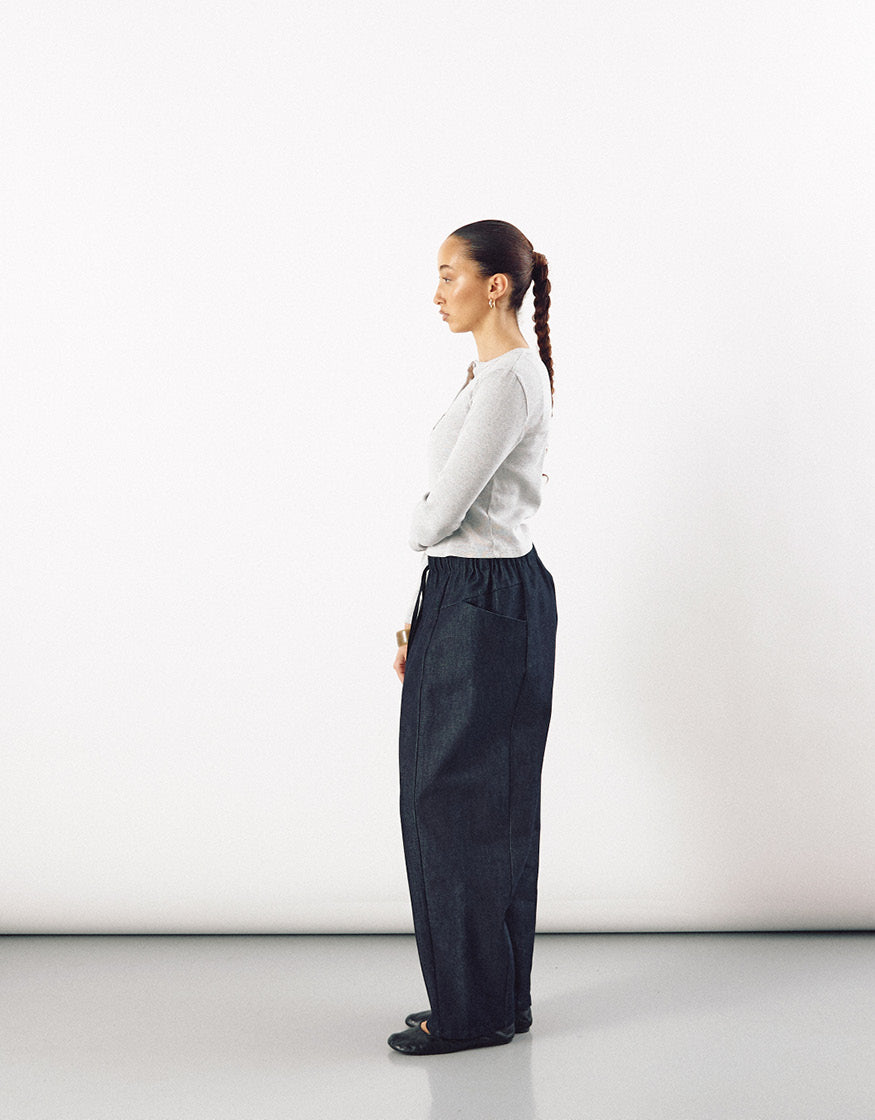 Profile view of a woman wearing indigo tapered pants paired with a light-colored fitted long-sleeve top, showcasing minimalist and timeless design with clean lines.
