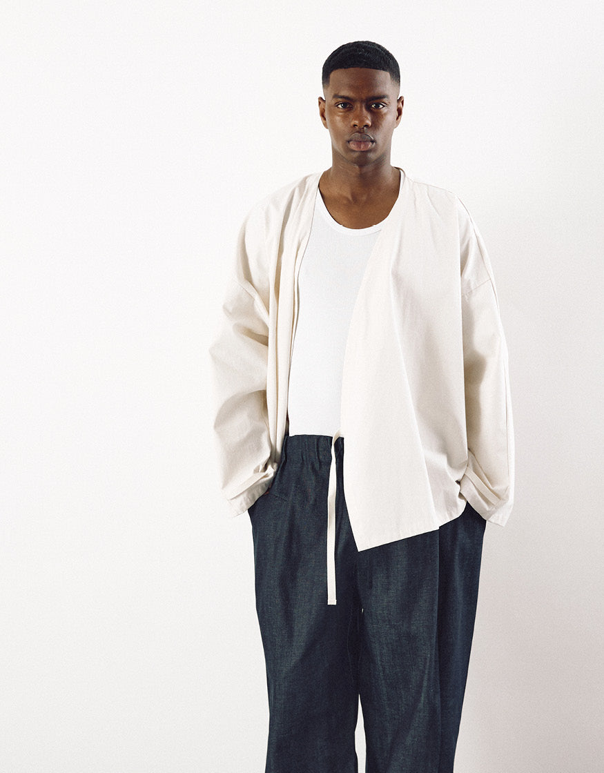 Male model wearing indigo tapered pants paired with a loose, cream-colored jacket and a white tank top, showcasing minimalist unisex fashion with clean lines and relaxed fit.