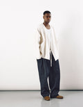 Male model wearing loose indigo tapered pants with a minimalist off-white open-front jacket and brown shoes against a plain white background, showcasing contemporary unisex design.