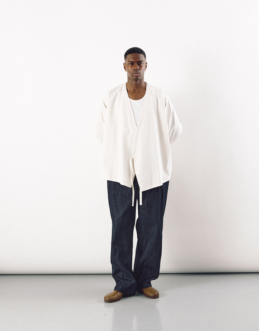 Male model wearing loose-fit indigo tapered pants paired with an oversized off-white top and brown shoes, showcasing minimalist unisex fashion with subtle asymmetry and volume.
