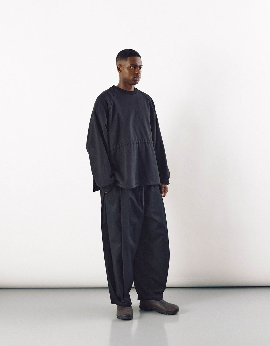 A man wearing loose-fitting black curved pants paired with a minimalist oversized black top, showcasing understated, contemporary unisex fashion with an emphasis on volume and movement.