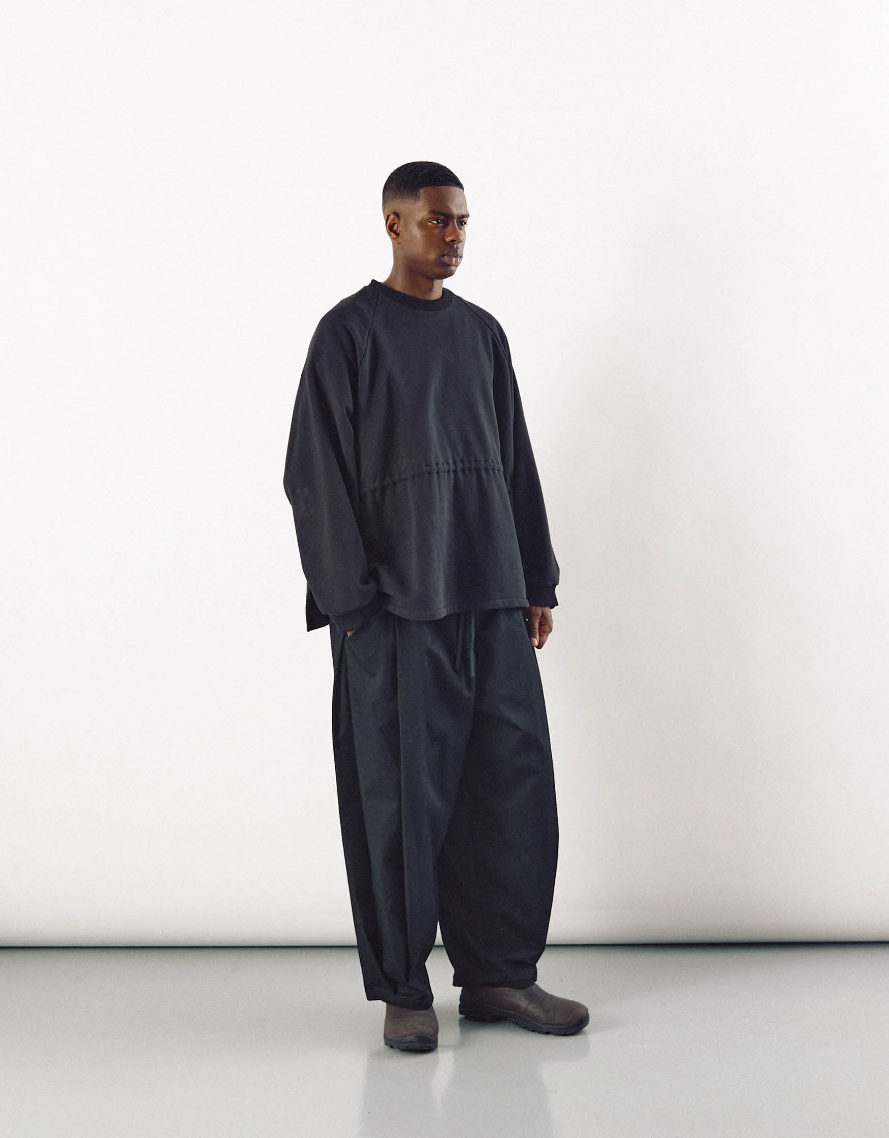 A man wearing loose-fitting black curved pants paired with a minimalist oversized black top, showcasing understated, contemporary unisex fashion with an emphasis on volume and movement.