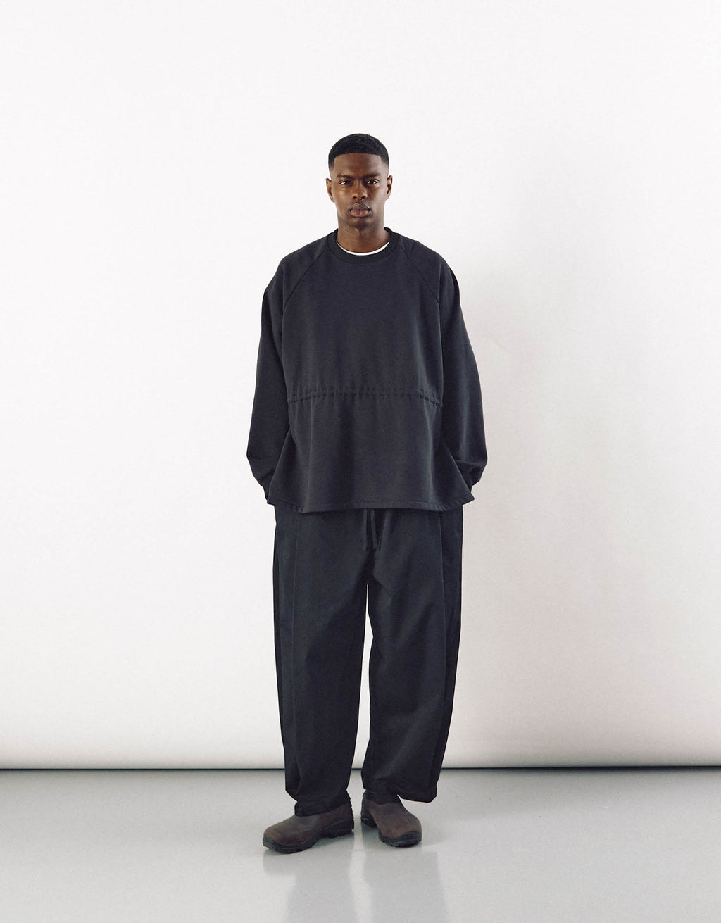 A male model wearing minimalist black curved pants paired with a loose-fitting black top, showcasing understated elegance and relaxed movement in a modern, unisex style.