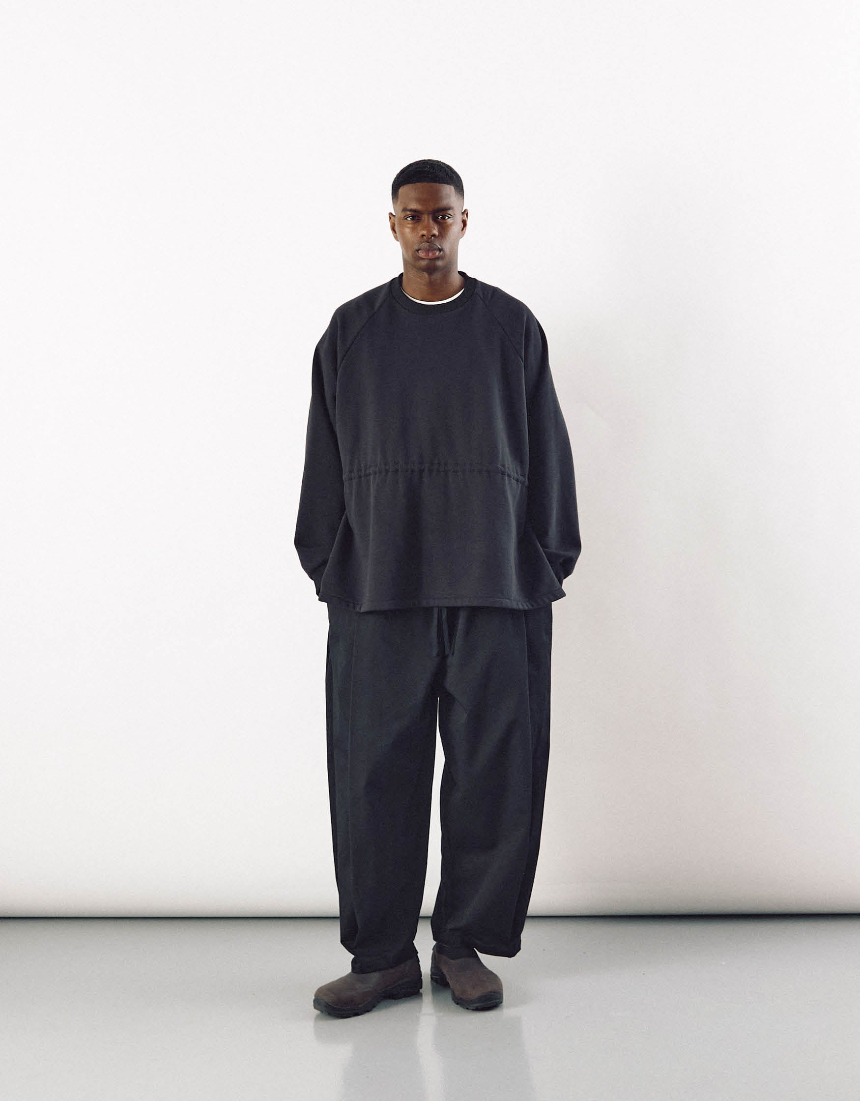 A male model wearing minimalist black curved pants paired with a loose-fitting black top, showcasing understated elegance and relaxed movement in a modern, unisex style.