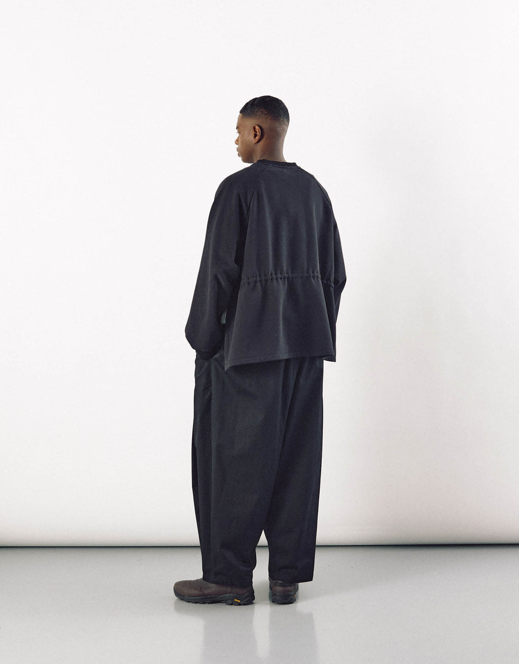 Model wearing loose-fitting black curved pants and a matching oversized jacket, showcasing minimalist, unisex design with emphasis on volume and freedom of movement.