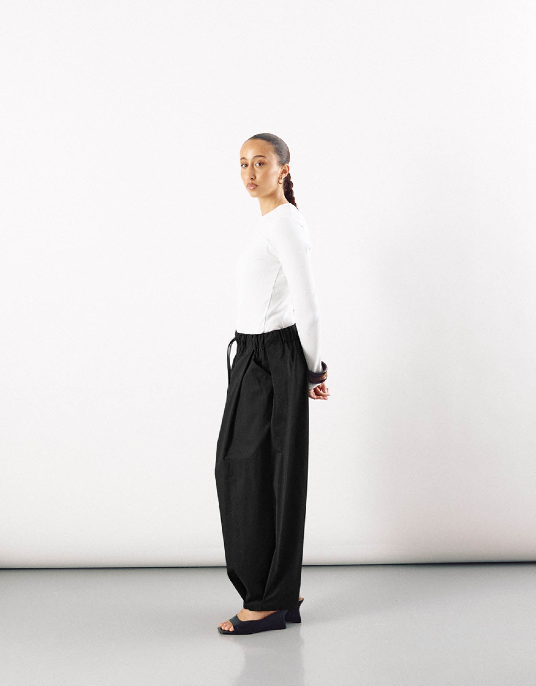Side profile of a model wearing minimalist black wide-leg pants paired with a white long-sleeve fitted top, emphasizing clean lines and subtle elegance.