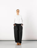 Woman wearing minimalist black curved pants with a relaxed fit and elastic waistband, paired with a simple white long-sleeve top and brown shoes, standing against a plain white background.