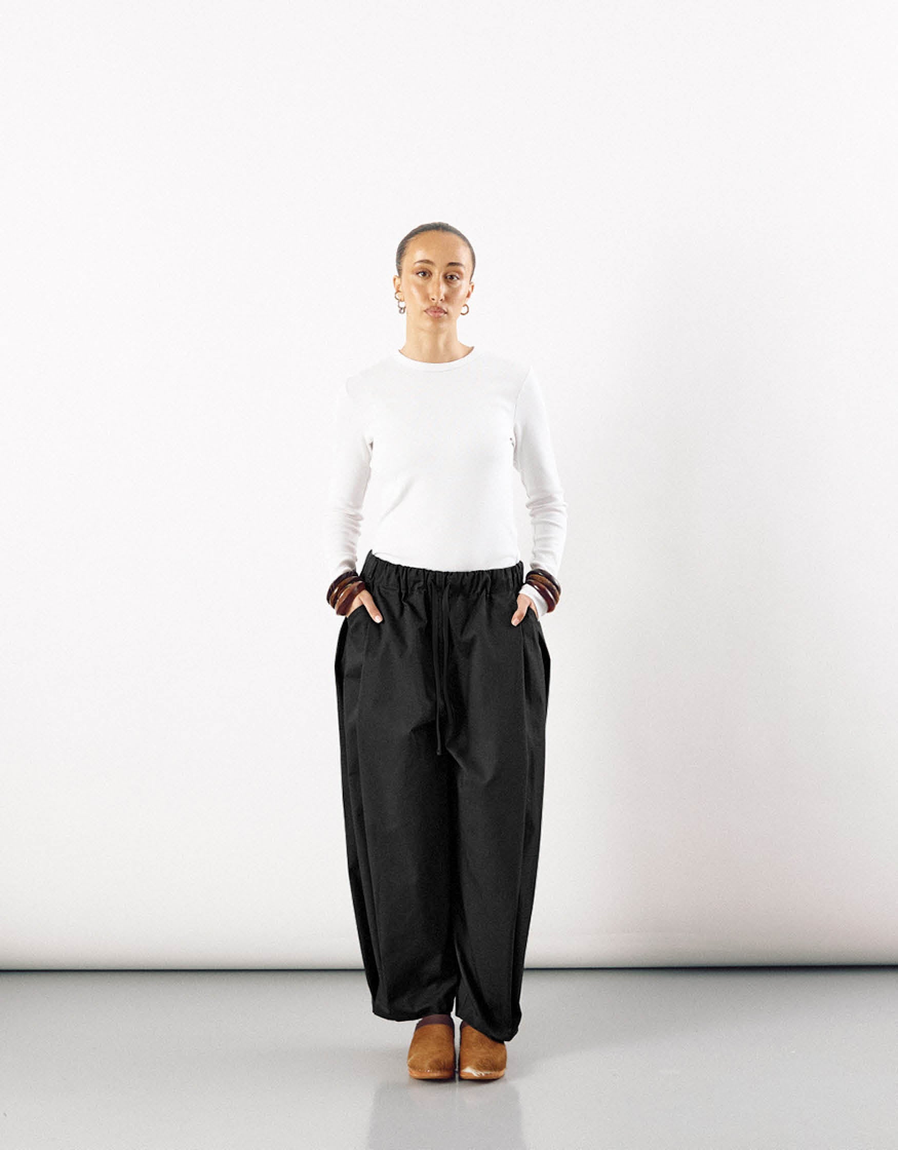Woman wearing minimalist black curved pants with a relaxed fit and elastic waistband, paired with a simple white long-sleeve top and brown shoes, standing against a plain white background.