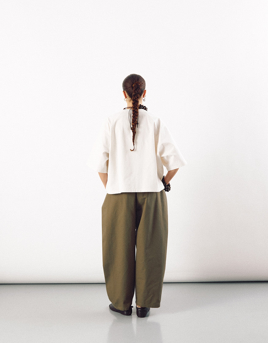 Rear view of a model wearing an off-white short sleeve shirt with a boxy, minimalist fit paired with loose, olive green wide-leg trousers against a plain white background.