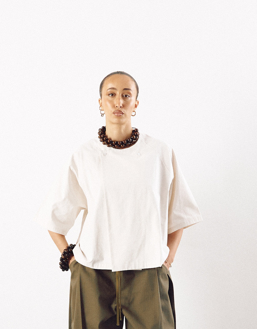 Model wearing an off-white minimalist short sleeve shirt with a boxy fit, paired with olive pants and accessorized with chunky dark bead jewelry, highlighting understated elegance.