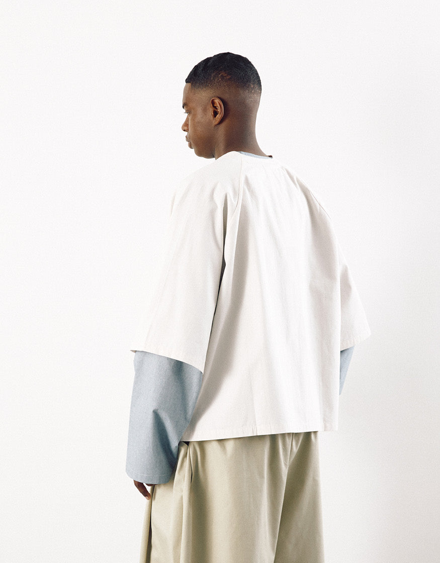 Man wearing a minimalist off-white short sleeve shirt layered over a long-sleeve garment, paired with loose beige trousers, showcasing contemporary design and understated elegance.