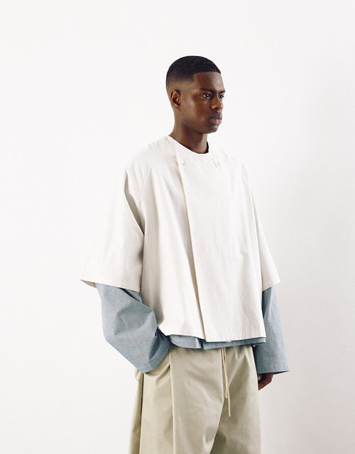 Male model wearing a minimalist off-white short sleeve shirt layered over a light blue long sleeve top, paired with beige trousers, showcasing contemporary unisex design with understated elegance.