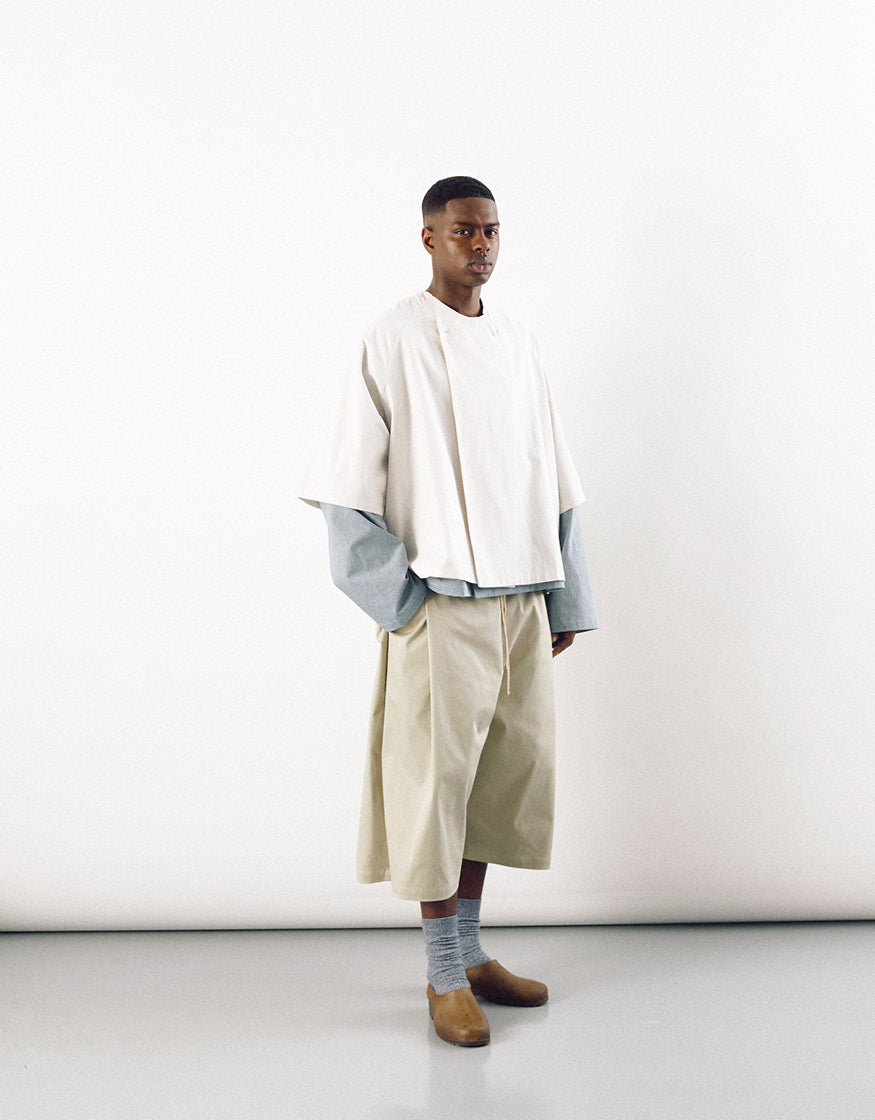 Male model wearing off-white short sleeve shirt layered over a long sleeve top with beige wide-leg cropped trousers and brown slip-on shoes, showcasing minimalist unisex fashion.