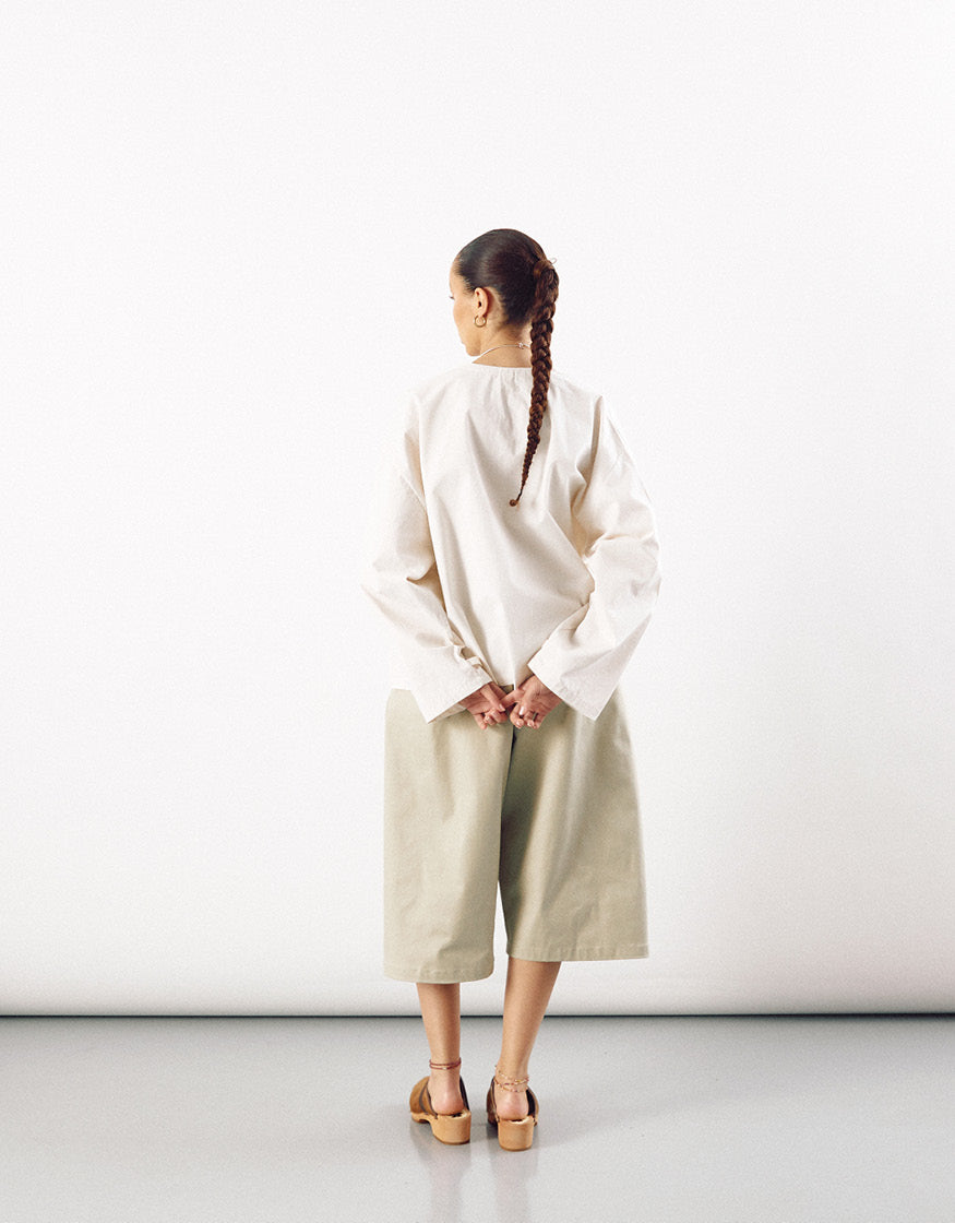 Rear view of a person wearing wide, straight-leg beige shorts paired with a loose, long-sleeve white top and brown sandals, standing against a plain light background.