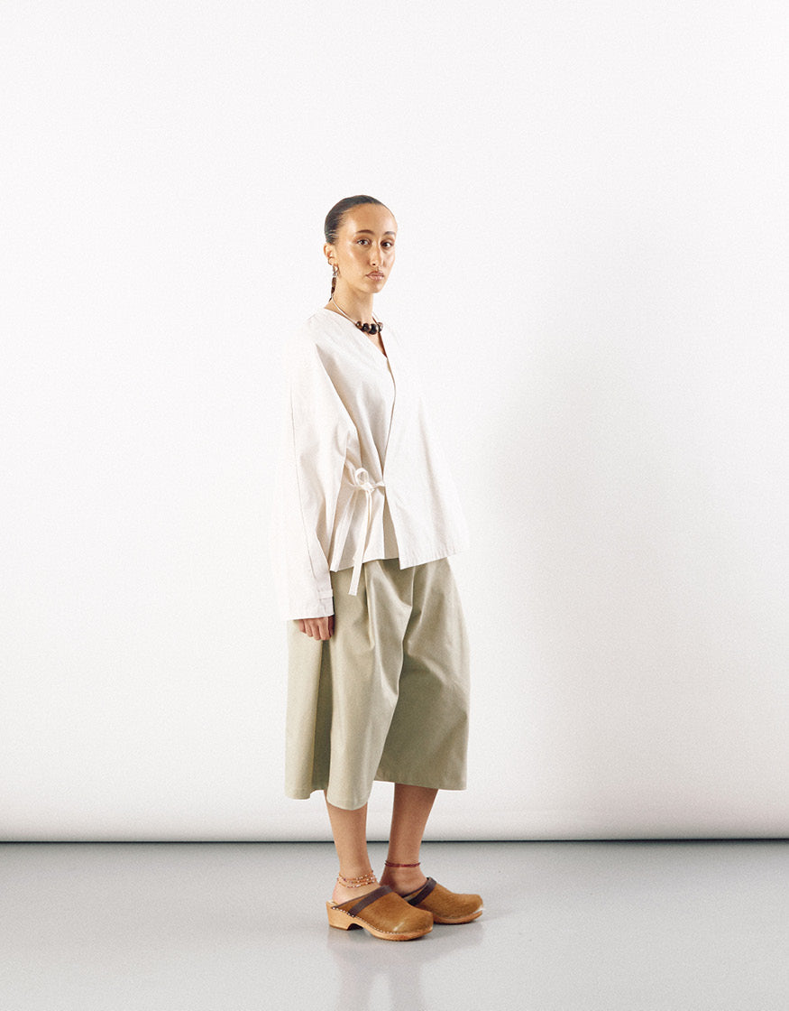 Model wearing beige wide straight-leg long shorts paired with a minimalist white wrap top and tan wooden clogs in a clean, contemporary setting.